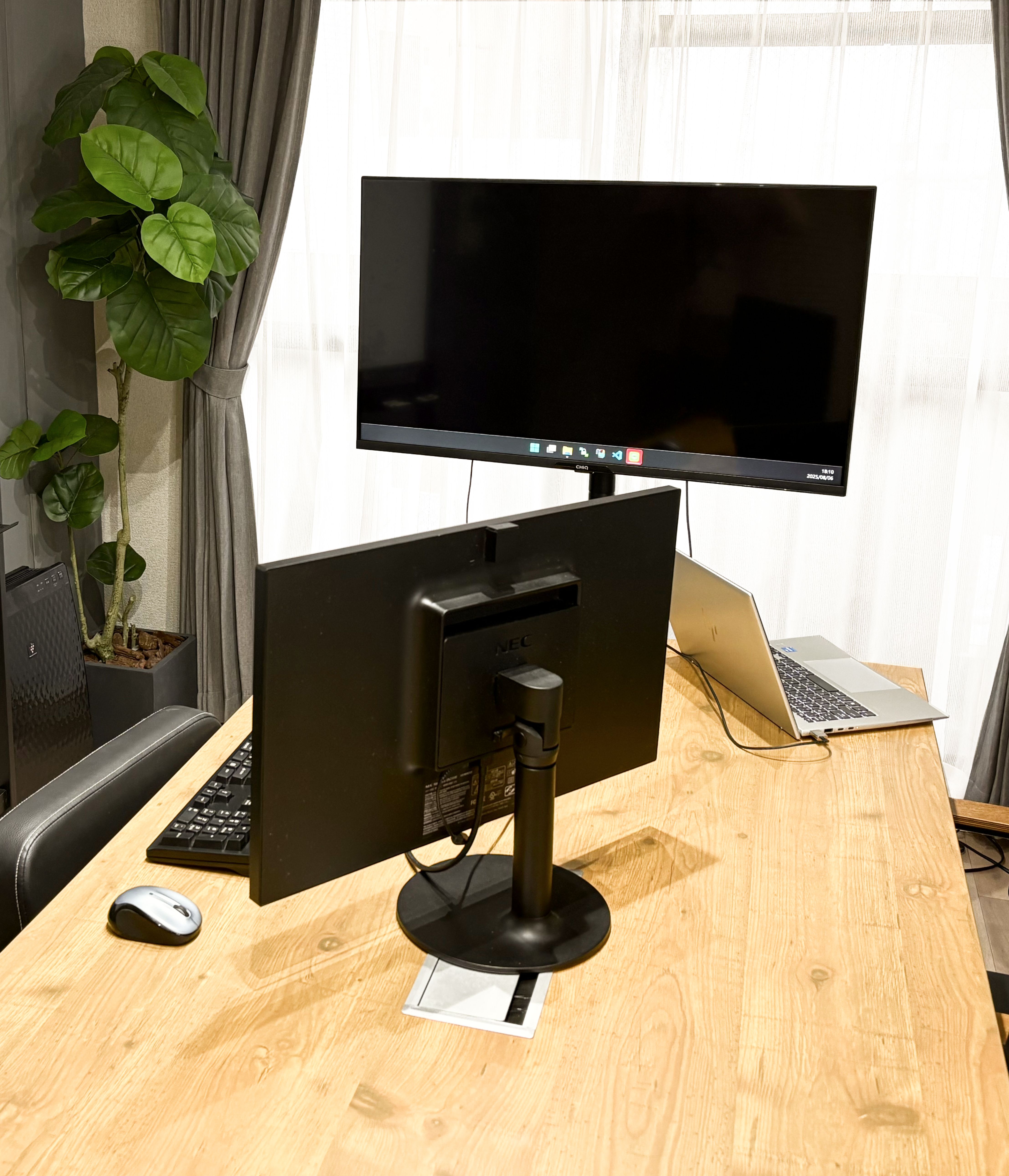 木目のテーブルの右側にノートパソコン、左側にディスプレイとキーボード、マウスが置いてある。奥には大きなモニターと観葉植物がある。