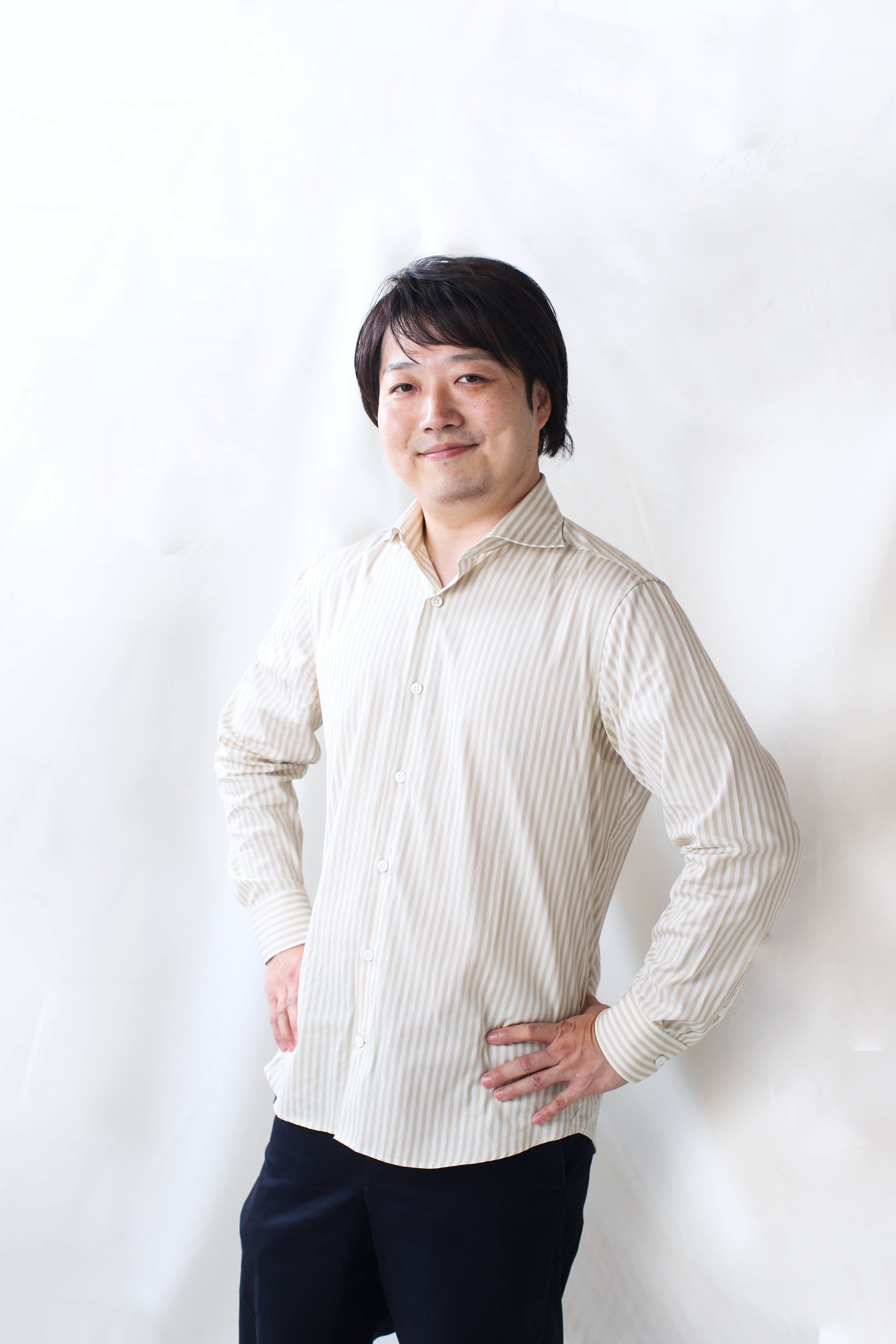 襟付きのベージュのストライプが入ったシャツを着た大西先生。柔和な笑顔で腰に手を置いて立っている。